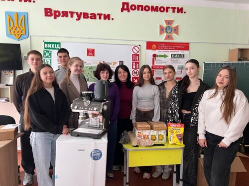 Group of students and a teacher pose around a coffee machine with snacks on a yellow cart in a Ukrainian classroom setting.
