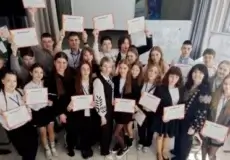 Large group of students in a classroom, smiling and holding certificates toward the camera, as if celebrating a ceremony.