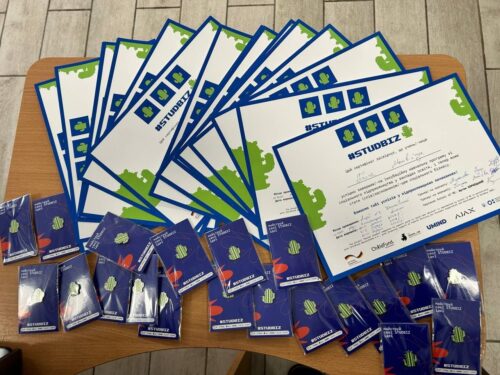 Fanned stack of certificates with blue borders and green map graphics arranged on a wooden table, accompanied by many blue event badges or cards at the bottom left and right edges.