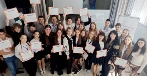 Large group of students in a classroom, smiling and holding certificates toward the camera, as if celebrating a ceremony.