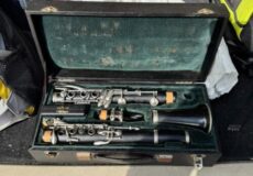 Open hard-shell case with a disassembled clarinet: mouthpiece, barrel, and three instrument sections laid inside with green lining.