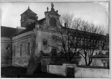 Black-and-white photo of an old stone church with twin towers and a surrounding wall, leafless trees nearby.