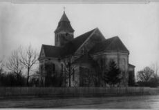 Stone church with a tall central tower and steep gabled roof, surrounded by leafless trees and a wooden fence.