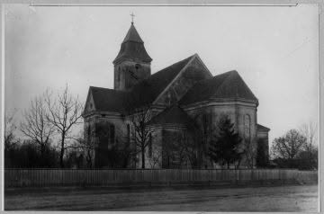 Stone church with a tall central tower and steep gabled roof, surrounded by leafless trees and a wooden fence.