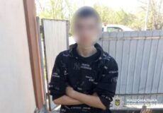 Person with a blurred face wearing a black hoodie with white text, standing with arms crossed outside near a fence on a sunny day (police watermark visible).