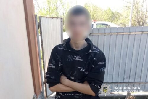 Person with a blurred face wearing a black hoodie with white text, standing with arms crossed outside near a fence on a sunny day (police watermark visible).