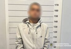 Blurred man in a light gray jacket stands before a height chart in a police mugshot, Ukrainian police logo visible.