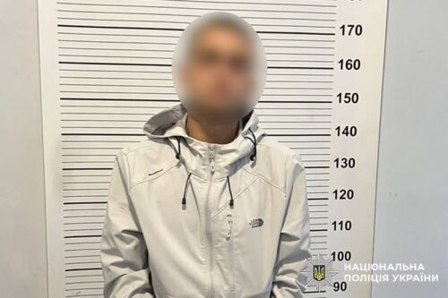Blurred man in a light gray jacket stands before a height chart in a police mugshot, Ukrainian police logo visible.
