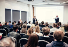 Audience in a conference room watching a presentation; two speakers stand at the front by a projected slide.
