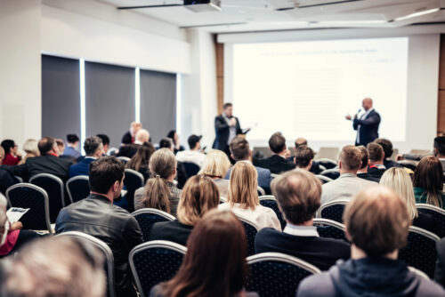 Audience in a conference room watching a presentation; two speakers stand at the front by a projected slide.