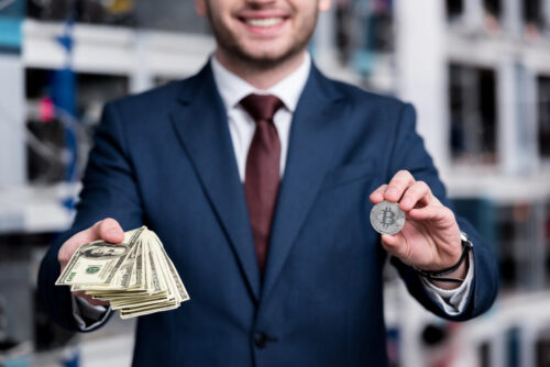 Smiling businessman in a blue suit holds a stack of cash in one hand and a Bitcoin coin in the other.