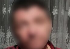 Person with a blurred face wearing a red plaid shirt, indoors in front of a floral lace curtain.