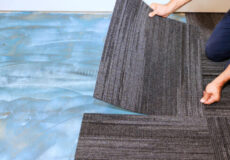 Person laying dark gray striped carpet tiles over a blue underlayment, aligning pieces on the floor.