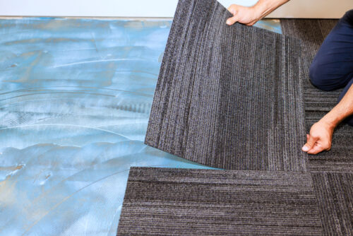 Person laying dark gray striped carpet tiles over a blue underlayment, aligning pieces on the floor.