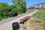 Bench beside a trash can along a curved path in a sunny park, with bushes and apartments in the background.