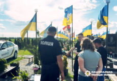 Police officer in a dark uniform at a memorial site, talking to visitors among blue and yellow Ukrainian flags.