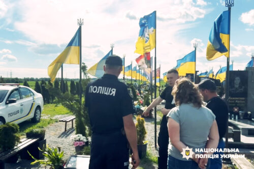 Police officer in a dark uniform at a memorial site, talking to visitors among blue and yellow Ukrainian flags.