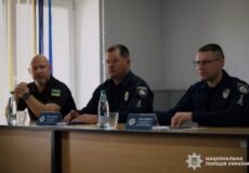 Three Ukrainian police officers sit at a panel table in a conference room, with water bottles and nameplates, the National Police of Ukraine emblem on their uniforms and a Ukrainian flag on the left sleeve visible.