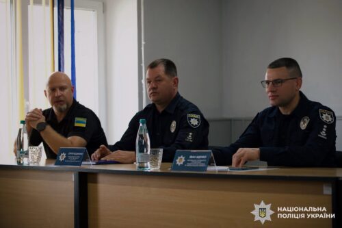 Three Ukrainian police officers sit at a panel table in a conference room, with water bottles and nameplates, the National Police of Ukraine emblem on their uniforms and a Ukrainian flag on the left sleeve visible.