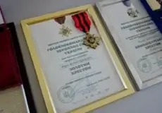 Three framed certificates with attached medals arranged on a surface; center frame has a gold border and red-and-black ribbon, flanked by a gold medal on the left and a blue ribbon on the right.