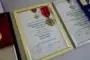 Three framed certificates with attached medals arranged on a surface; center frame has a gold border and red-and-black ribbon, flanked by a gold medal on the left and a blue ribbon on the right.