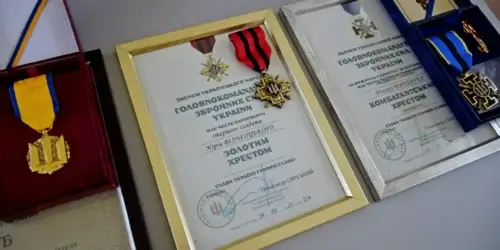 Three framed certificates with attached medals arranged on a surface; center frame has a gold border and red-and-black ribbon, flanked by a gold medal on the left and a blue ribbon on the right.