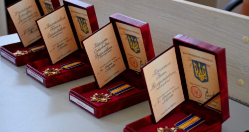 Row of open red boxes displaying medals with ribbons and certificates on a light table surface, arranged in a line.
