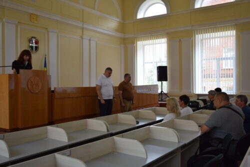 Woman at a podium addressing a small audience in a bright council chamber; two men stand toward the front as attendees listen.