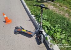 Electric kick scooter tipped over on a paved road beside a curb, with an orange traffic cone nearby and grass along the roadside; Ukrainian police watermark in the corner.