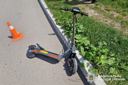 Electric kick scooter tipped over on a paved road beside a curb, with an orange traffic cone nearby and grass along the roadside; Ukrainian police watermark in the corner.