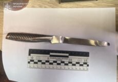 Evidence photo: metal knife with ribbed handle laid on white sheet beside a scale ruler; watermark from Ukrainian Prosecutor's Office in corner.