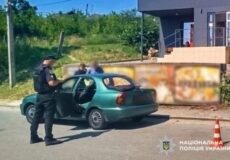 Policeman in dark uniform by a green car with its door open on a street; blurred passenger nearby, building and orange cone in background.