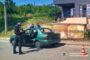 Policeman in dark uniform by a green car with its door open on a street; blurred passenger nearby, building and orange cone in background.