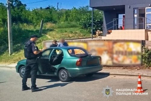 Policeman in dark uniform by a green car with its door open on a street; blurred passenger nearby, building and orange cone in background.