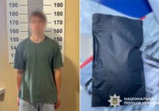 Blurred man in a green shirt stands before a height chart at a police station; to his right is a sealed black evidence bag with Ukrainian police emblem.
