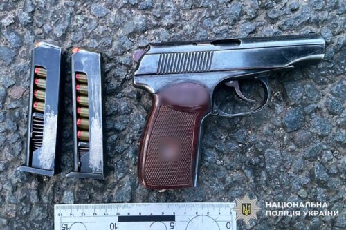 Semi-automatic pistol with a brown textured grip, two loaded magazines, on rough asphalt, with a ruler for scale and a National Police of Ukraine watermark.