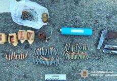 Assorted ammunition and firearm parts neatly arranged on pavement, including bullets in rows, a pistol with magazines, and a plastic bag of rounds; National Police of Ukraine logo in the corner.