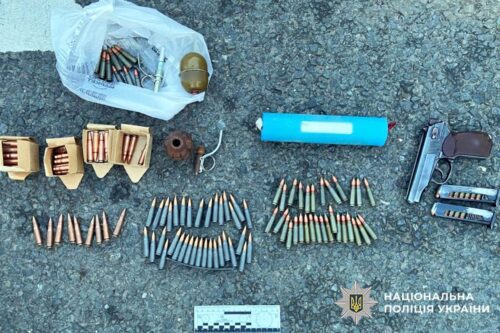 Assorted ammunition and firearm parts neatly arranged on pavement, including bullets in rows, a pistol with magazines, and a plastic bag of rounds; National Police of Ukraine logo in the corner.