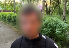 Person with a blurred face standing on a park path, wearing a black Nike jacket and backpack, with a police watermark in the corner.
