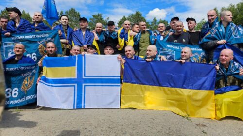 Group of supporters outdoors, wearing blue and yellow clothing, holding a Ukrainian flag and a Finnish flag in front of a blue sky.