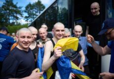 Group of bald men in athletic wear disembarking from a bus, one in the front holding a blue-and-yellow flag