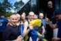 Group of bald men in athletic wear disembarking from a bus, one in the front holding a blue-and-yellow flag