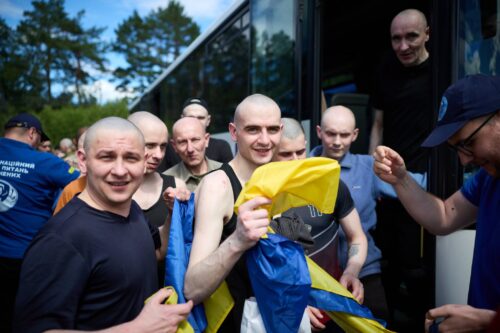 Group of bald men in athletic wear disembarking from a bus, one in the front holding a blue-and-yellow flag