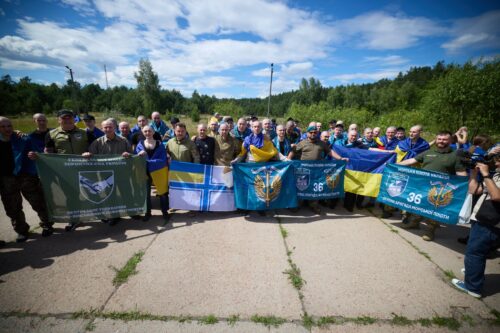Group of people standing in a line outdoors on a sunny day, holding blue-and-yellow Ukrainian flags and banners with emblems, suggesting a memorial or veterans gathering.