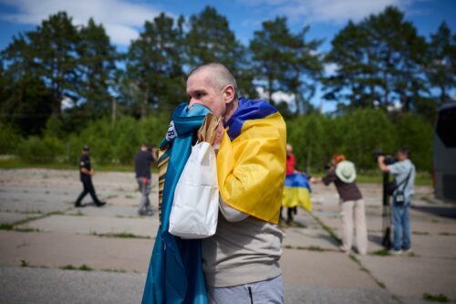 Man wrapped in a Ukrainian flag on an airfield, preparing or resting, with people and trees in the background under a blue sky.
