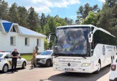 Large white tour bus with passengers visible through the windshield, parked near a police car and a small white building with a blue roof amid trees on a sunny day.