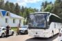 Large white tour bus with passengers visible through the windshield, parked near a police car and a small white building with a blue roof amid trees on a sunny day.