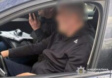 Two men sit in a car; one driver, one passenger with a camera and papers; passenger's face blurred; Ukrainian police emblem in the corner.