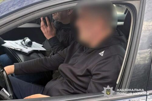 Two men sit in a car; one driver, one passenger with a camera and papers; passenger's face blurred; Ukrainian police emblem in the corner.