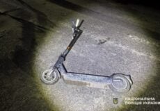 Electric scooter lying on its side on a dimly lit road at night, with the National Police of Ukraine logo in the corner.
